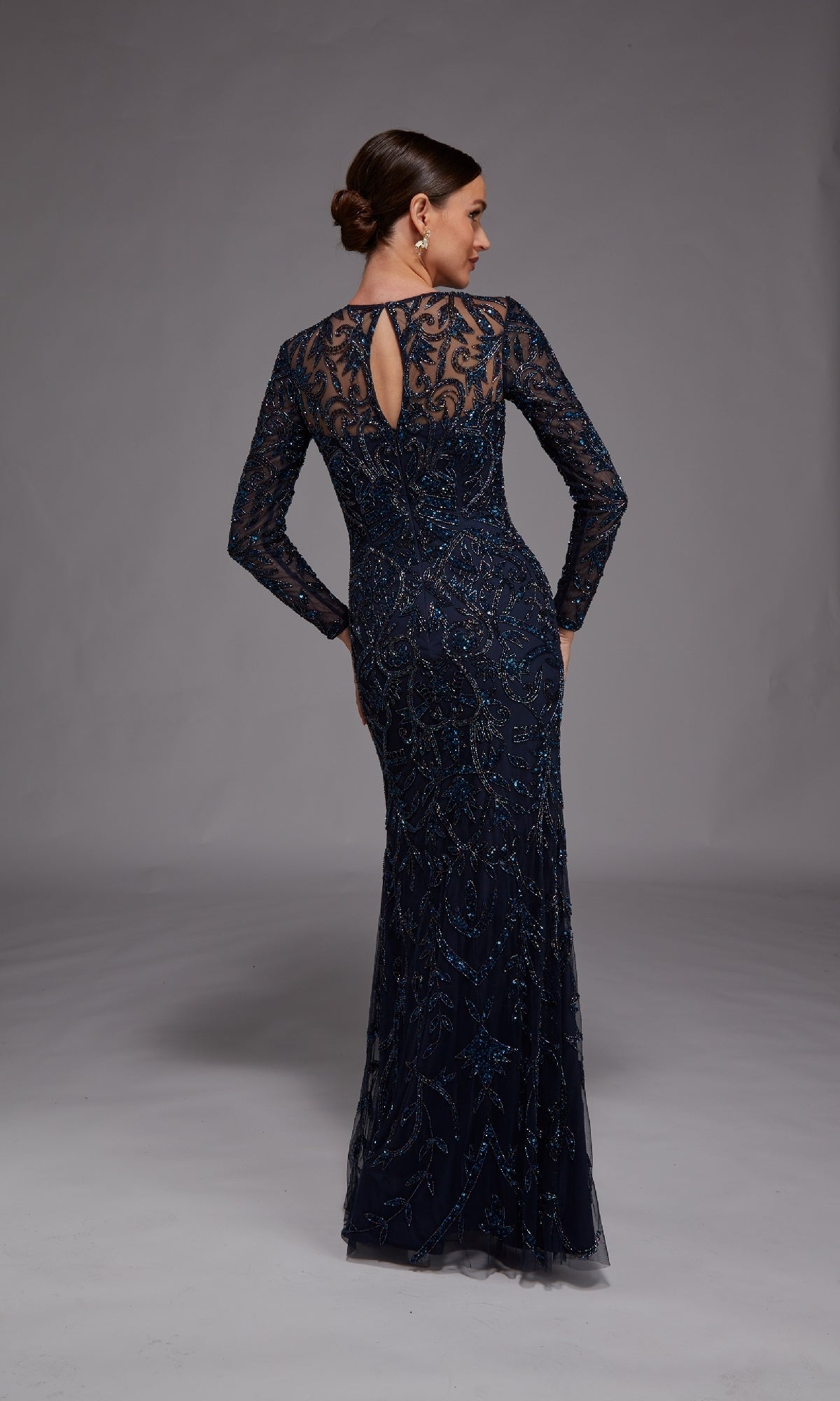 A woman models the Alyce 27778, a dark blue long formal dress with intricate beading—an elegant choice for the mother of the bride—standing with her back to the camera against a plain grey background.